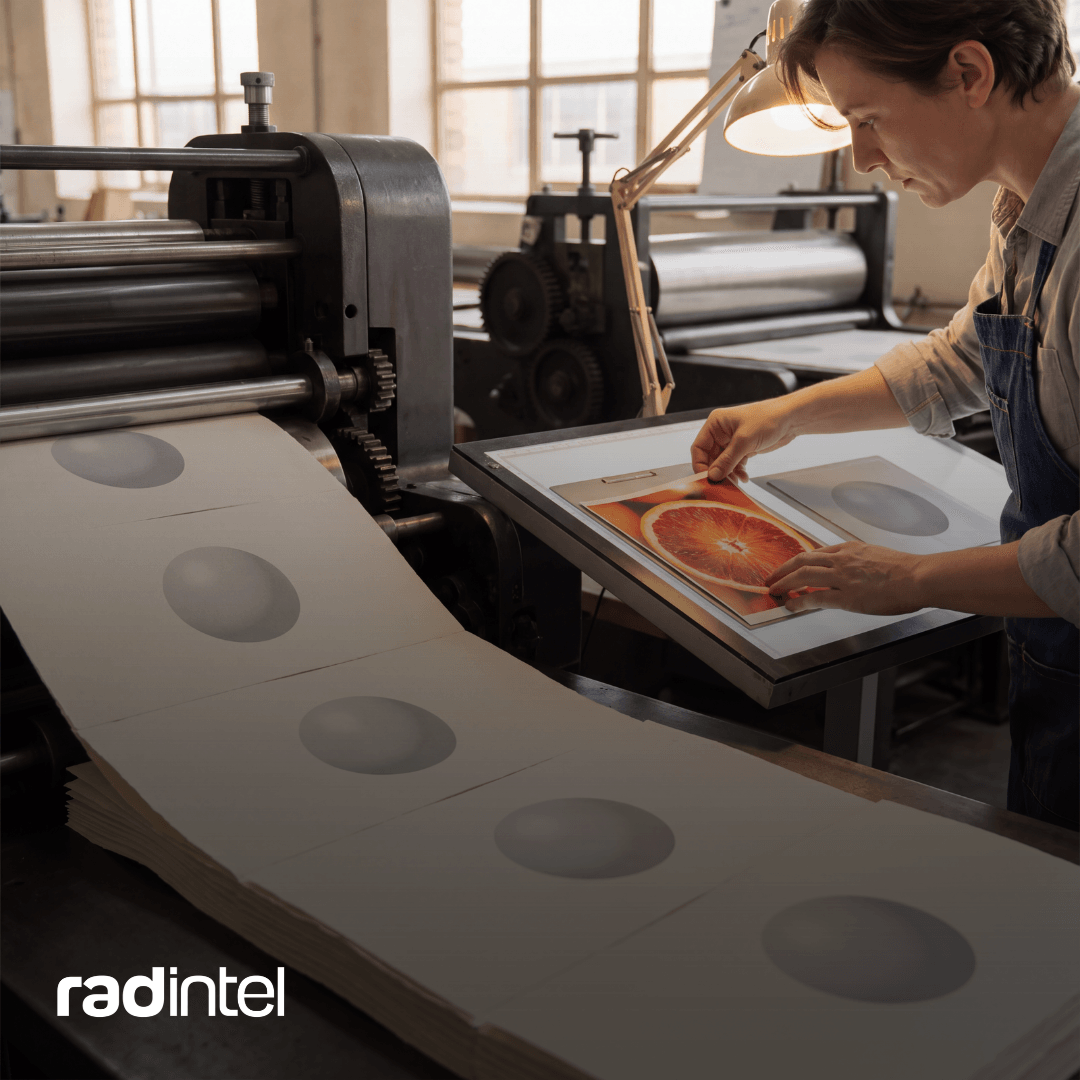 A printing press turns out a row of identical sheets, each printed with the same gray circle, while a printmaker examines a vivid blood orange print on a light table.
